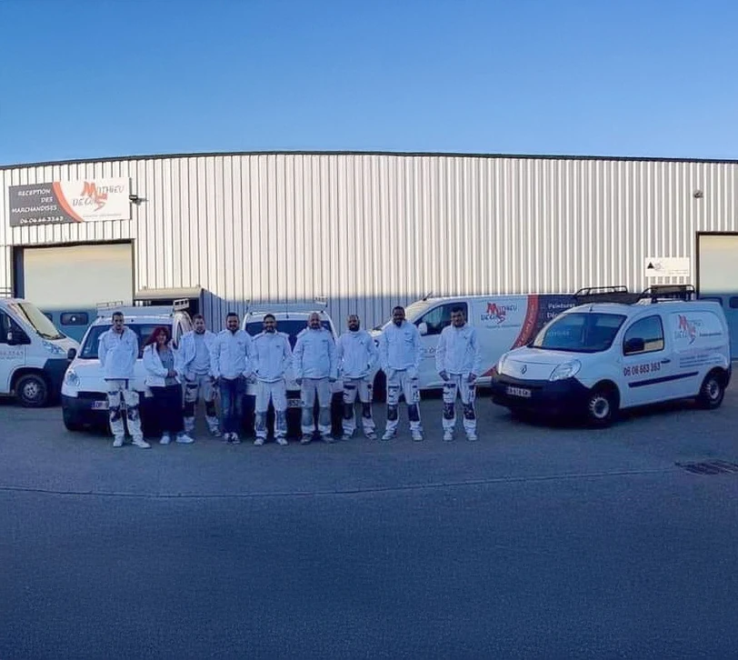 Équipe de peintres professionnels en combinaisons blanches posant devant leur flotte de véhicules utilitaires blancs sur parking d'entreprise