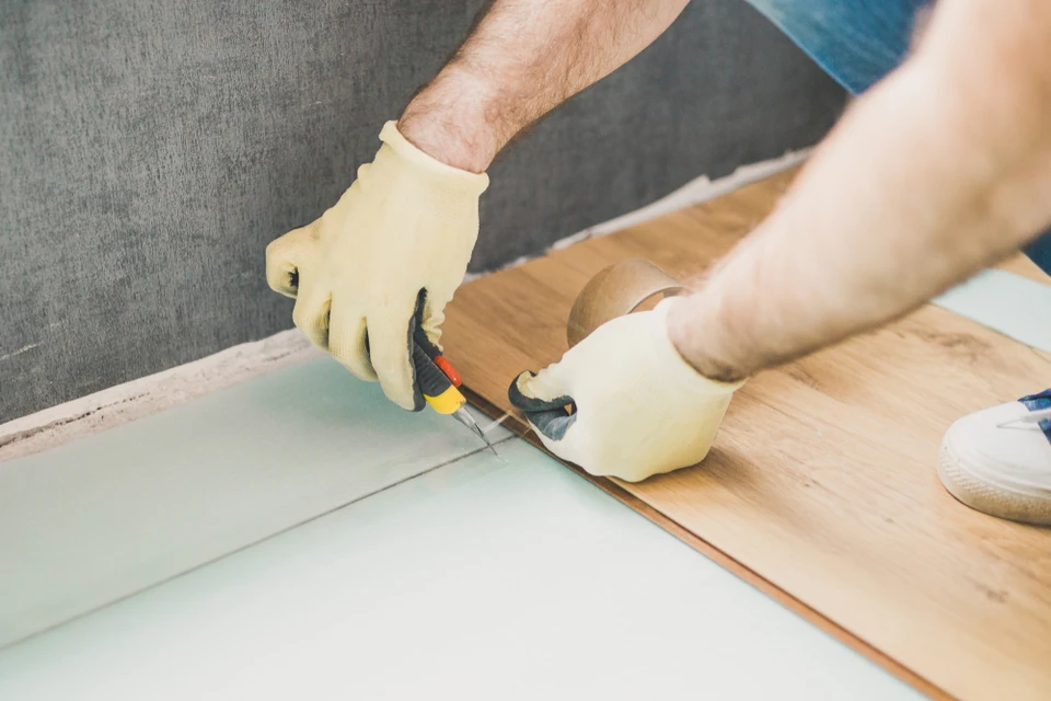 Intérieur de chantier, ouvrier en gants de protection jaunes utilisant une scie sauteuse électrique pour découper des lames de parquet stratifié